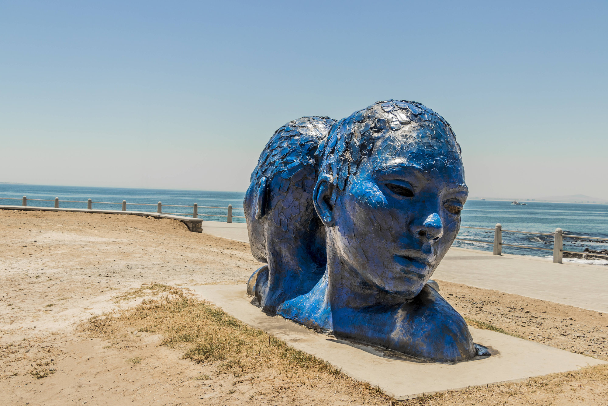 Kunstinstallation: zwei voneinander abgewandte menschliche Köpfe aus tiefblauem, glänzendem Material. Sie stehen auf einer Sandfläche an einer Küste. Im Hintergrund ist ein weißes Geländer, dahinter hellblaues Wasser.