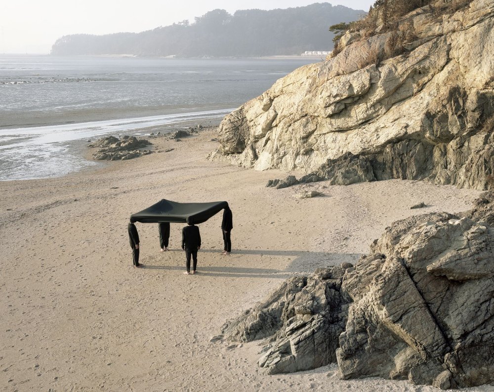 Vier ganz in schwarz gekleidete Personen führen an einem Strand vor Felsklippen eine Performance durch, bei denen ihre Köpfe mit einem viereckigen schwarzen Tuch verbunden sind.
Bild: Seok-Woo Song