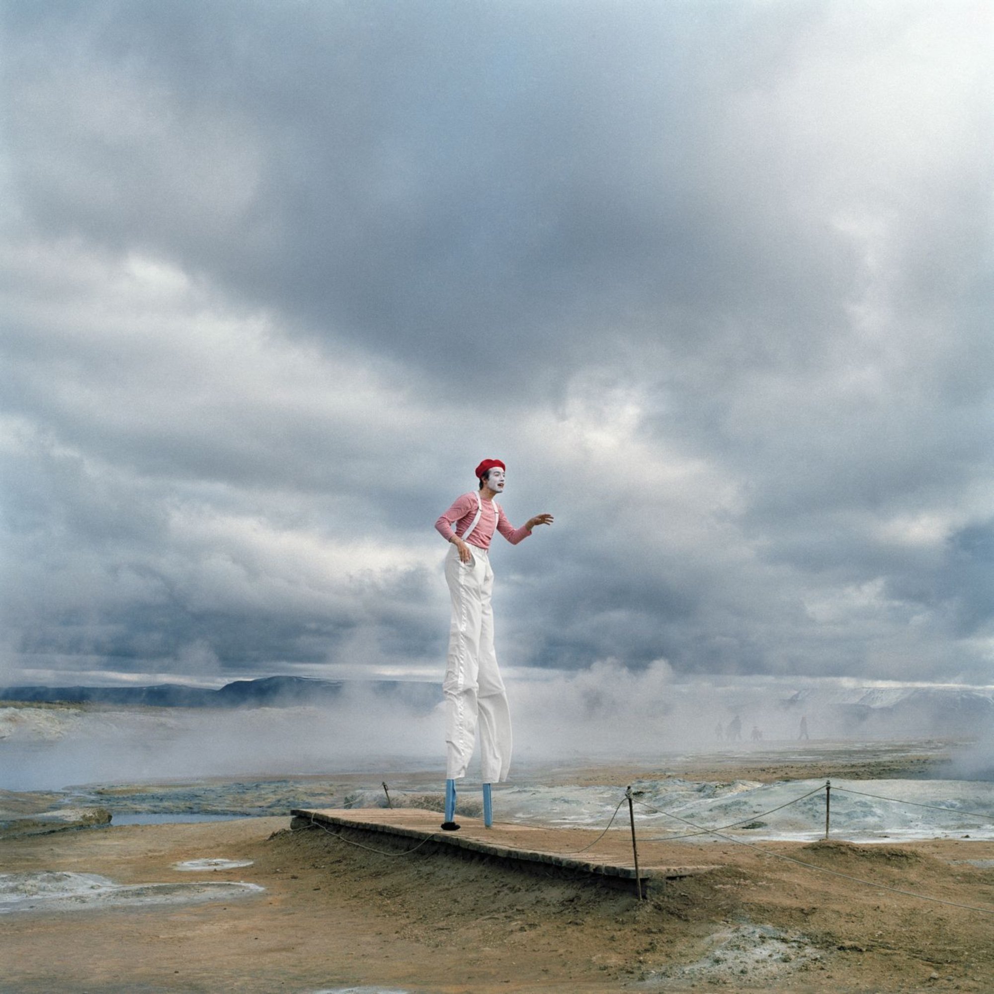 Ein weißgekleideter, geschminkter Pantomime auf Stelzen auf einem Steg am Strand vor wolkenverhangenem Himmel
Bild: Johanna Maria Fritz | Agentur Ostkreuz